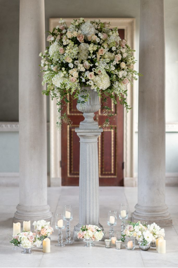 Urn flower arrangement with blush and pink flowers, standing on concrete plinth with bud vases and candles at the base
