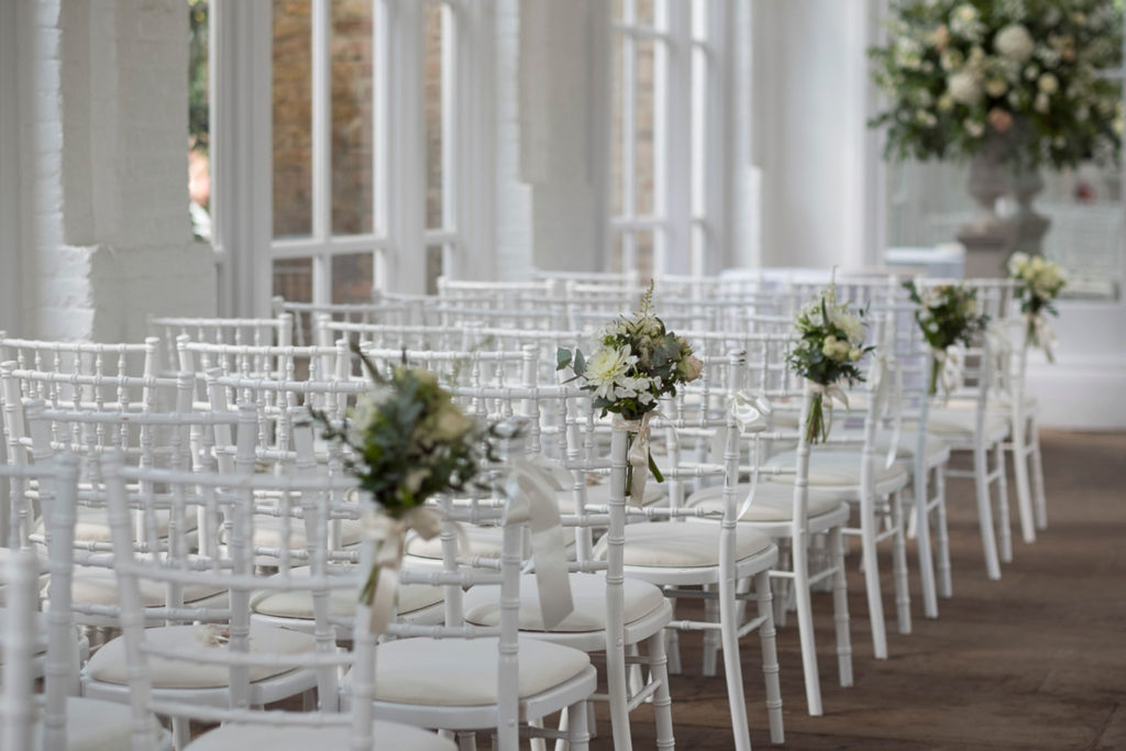 Wedding ceremony with Chiavari chairs, pink flower posie on shoulder at Holland Park Orangery