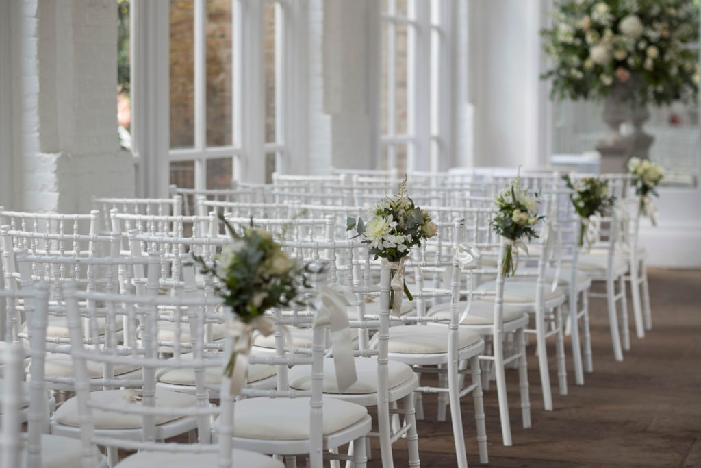 Wedding ceremony with Chiavari chairs, pink flower posie on shoulder at Holland Park Orangery