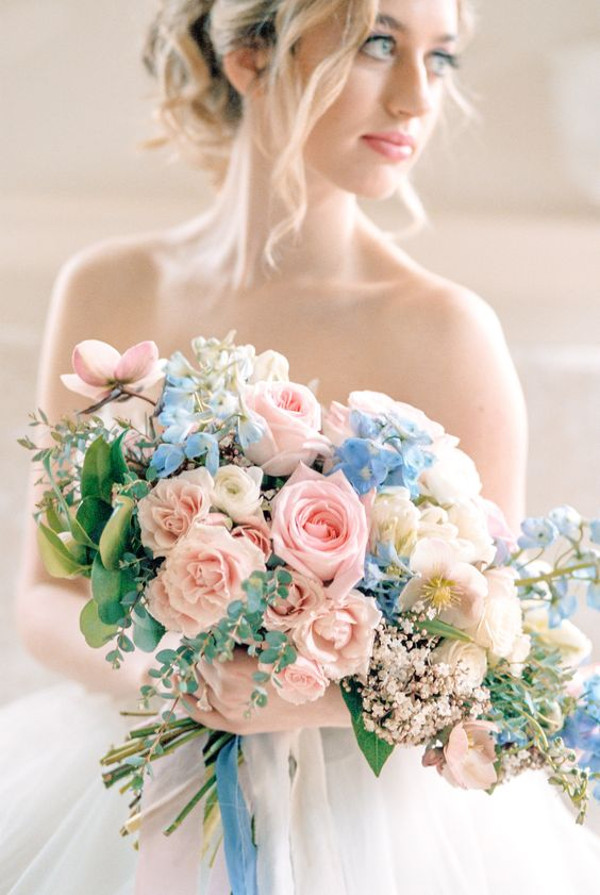 Bride holding a bouquet of pink and blush roses, blue delphiniums and greenery, with a blue ribbon